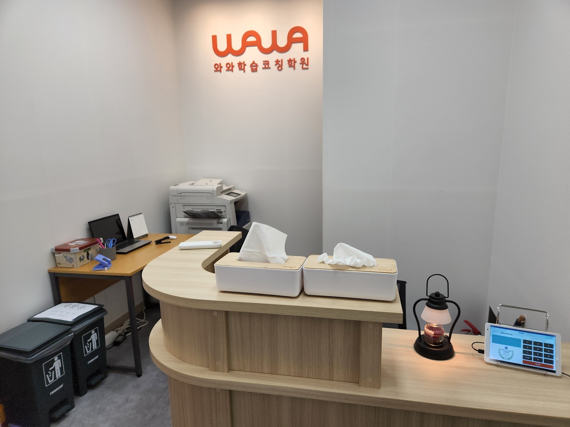 범박점 안내 데스크 — 흰 벽 위쪽에 주황색 WAWA 로고와 와와학습코칭학원 글자가 붙어 있고, 그 아래 원목 곡선 데스크가 놓여 있다. 데스크 위에 티슈 상자 두 개와 스탠드 조명, 출결 확인용 태블릿이 있고, 뒤쪽으로 복합기와 노트북이 놓인 업무용 책상이 보인다