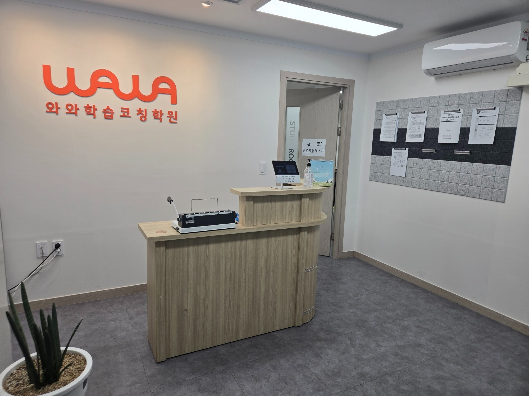 와와학습코칭 사직점 안내 데스크 — 흰 벽의 주황색 WAWA 와와학습코칭학원 로고, 출결 확인용 태블릿이 놓인 나무색 데스크, STUDY ROOM 문에 붙은 출결 확인 안내문, 오른쪽 게시판의 입시 안내 자료
