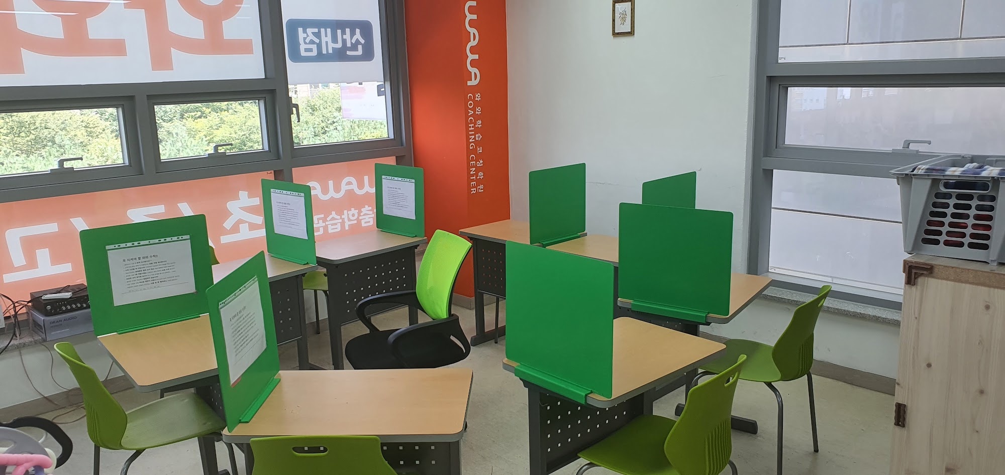 산내점 학습실 — 나무색 책상마다 초록색 가림판이 세워져 있고 가림판에는 학습 안내가 담긴 종이가 끼워져 있다. 연두색 의자가 책상마다 놓여 있으며, 안쪽 벽에는 와와 코칭센터 로고가 세로로 적힌 주황색 배너가 서 있고 창밖으로 나무가 보인다