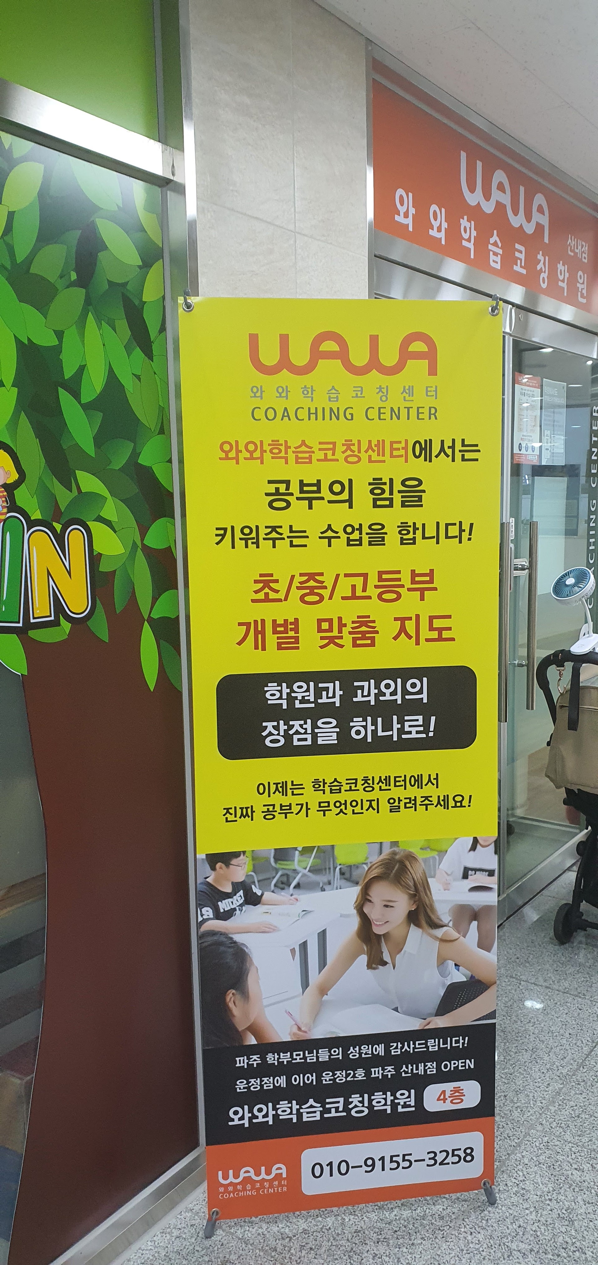 산내점 입구 앞에 세워진 안내 배너 — 노란 바탕에 와와학습코칭센터 로고와 '공부의 힘을 키워주는 수업을 합니다', '초/중/고등부 개별 맞춤 지도'라는 문구가 적혀 있다. 배너 아래에는 '운정점에 이어 운정2호 파주 산내점 OPEN, 와와학습코칭학원 4층'이라고 쓰여 있고, 뒤쪽 유리문 위에 주황색 산내점 간판이 보인다