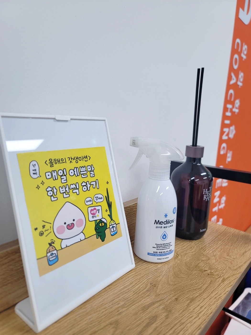 옥정점 데스크 위에 놓인 작은 액자 — 노란 바탕에 '올해의 갓생미션, 매일 예쁜말 한 번씩 하기'라는 문구와 캐릭터 그림이 그려져 있다. 옆에는 소독제와 디퓨저가 놓여 있고 뒤쪽에 코칭센터 안내판이 보인다
