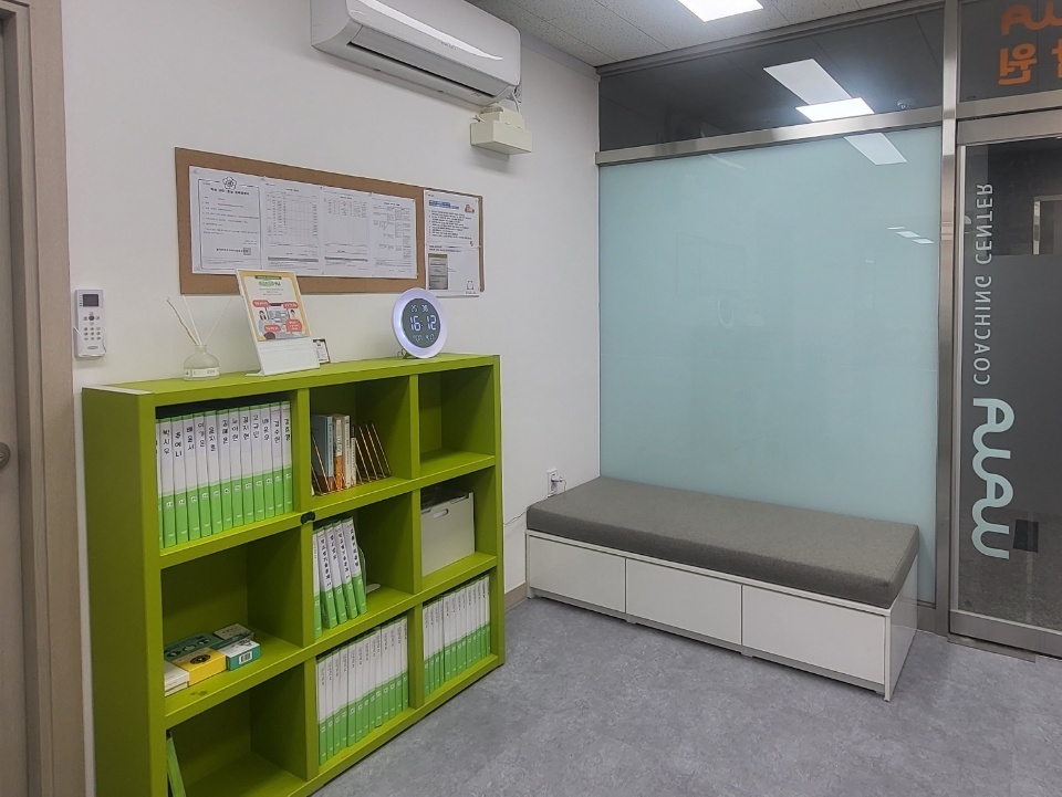 운정호수점 대기 공간 — 연두색 칸 책장에 초록색 파일철과 교재가 종류별로 꽂혀 있고, 위쪽 벽에는 안내문을 붙인 게시판과 둥근 벽시계가 걸려 있다. 오른쪽에는 앉아서 기다릴 수 있는 긴 벤치와 wawa COACHING CENTER 로고가 들어간 유리문이 있다