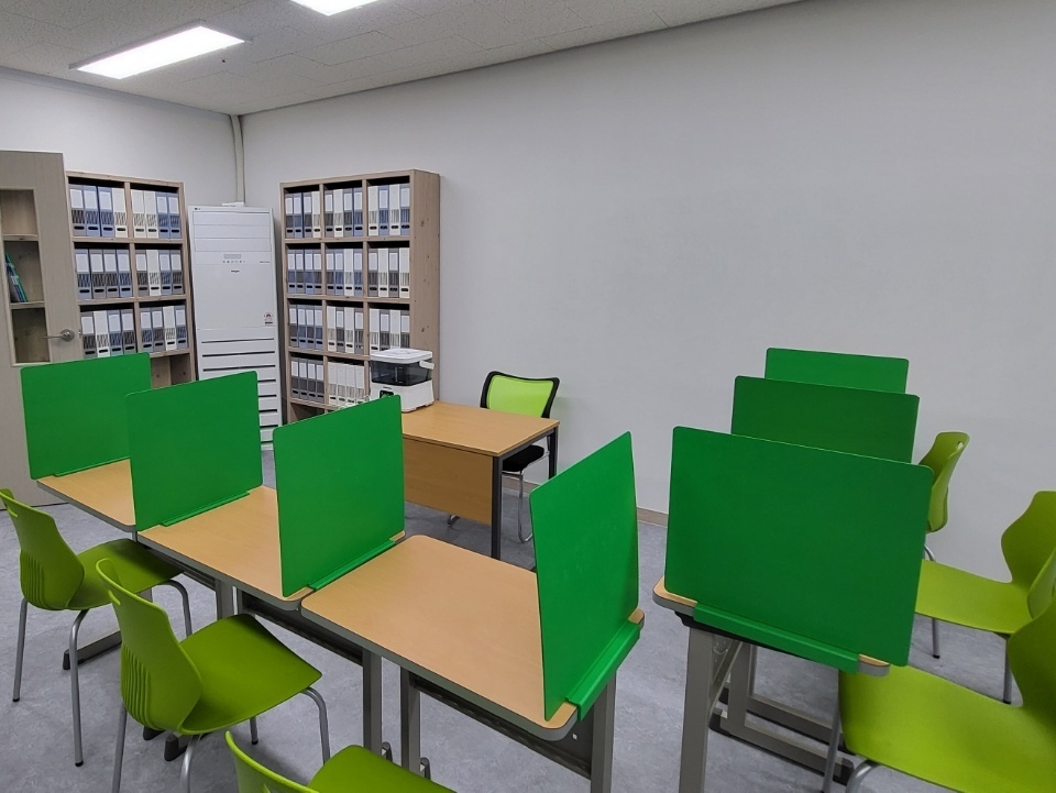 운정호수점 학습실 — 초록색 칸막이를 세운 책상 여러 개가 두 줄로 놓여 있고 연두색 의자가 딸려 있다. 뒤쪽 벽면에는 흰색 바인더가 가득 꽂힌 원목 선반 두 개와 프린터, 선생님 책상이 있다