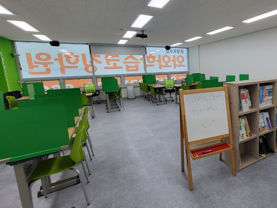 운정호수점 교실 — 창가 전체에 내린 블라인드에 와와학습코칭센터 글자가 크게 인쇄되어 있고, 왼쪽으로 초록색 칸막이를 세운 책상들이 줄지어 놓여 있다. 천장에는 프로젝터가 달려 있고, 오른쪽에는 책이 가득한 원목 칸 책장과 이젤형 화이트보드가 놓여 있다. 화이트보드에는 손글씨와 웃는 얼굴이 그려져 있다