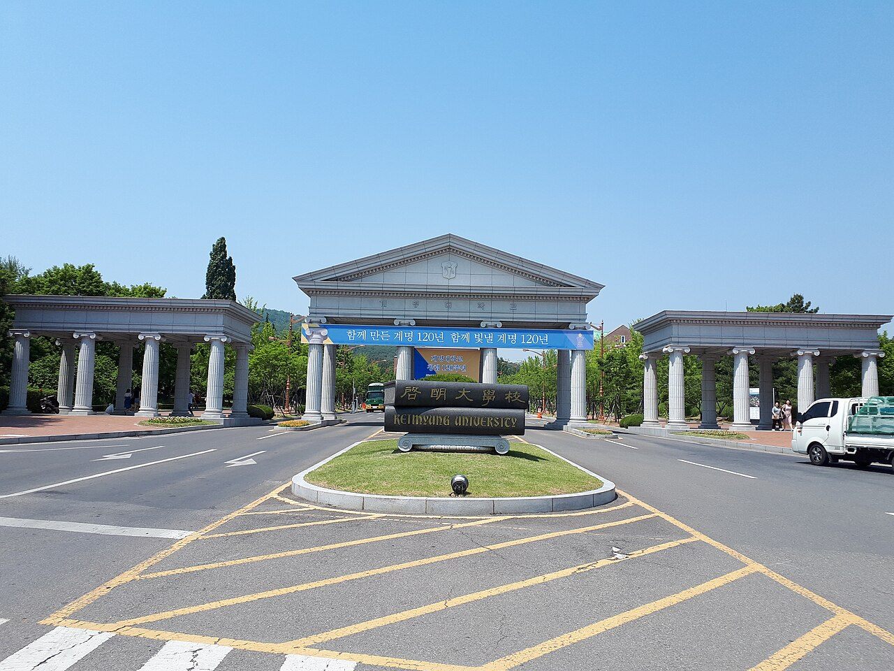 계명대학교 정문 — 그리스 신전풍의 흰 기둥 문 세 개가 나란히 서 있고, 가운데 도로 교통섬에 한자와 영문으로 계명대학교라고 새긴 검은 돌 표지석이 놓여 있다