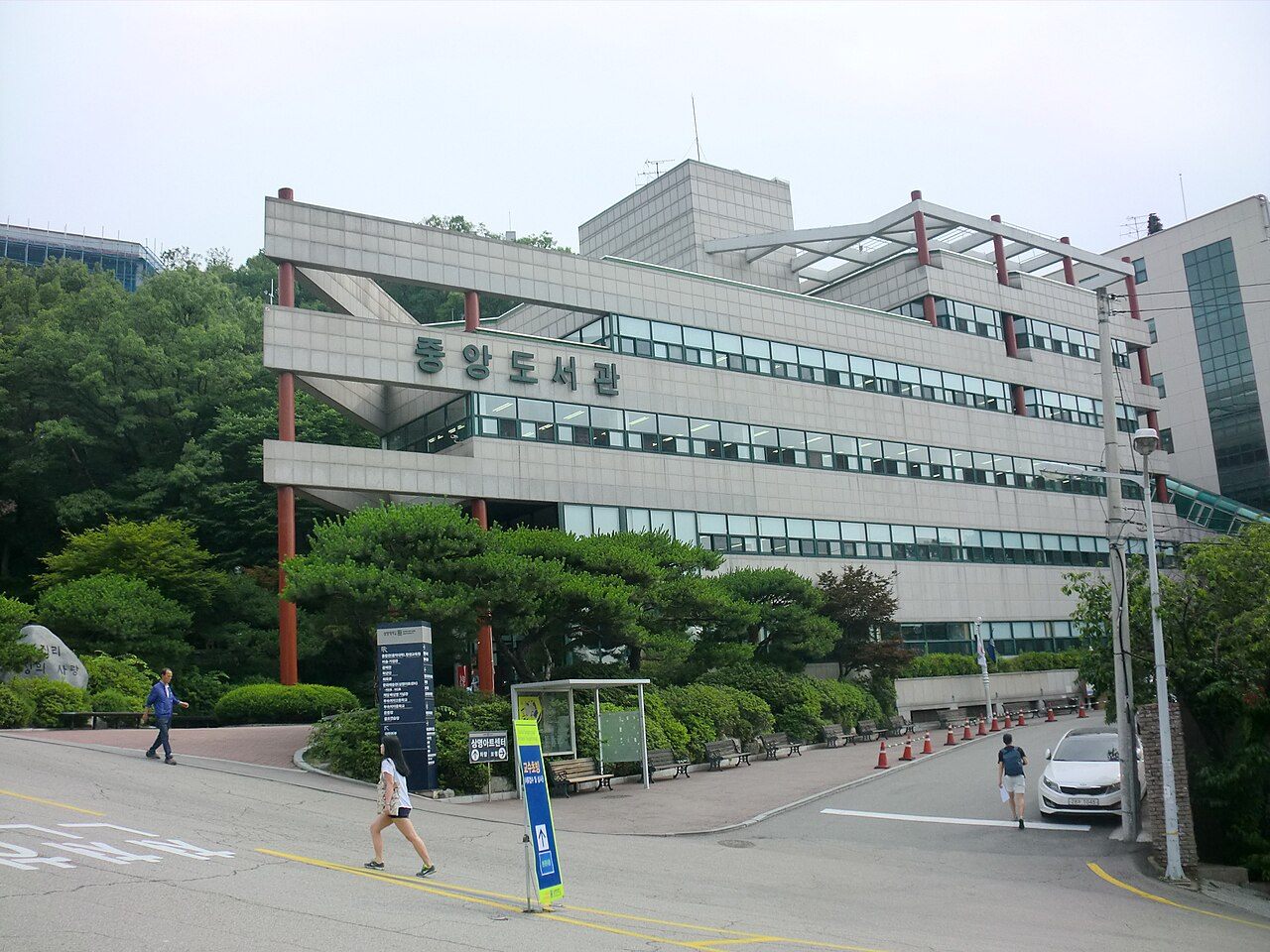 상명대학교 서울캠퍼스 중앙도서관 — 언덕을 따라 계단식으로 올라가는 회색 콘크리트 건물에 붉은 기둥이 세로로 서 있고, 건물 정면 위쪽에 중앙도서관이라는 큰 글자가 붙어 있다. 건물 앞으로는 소나무와 관목이 심긴 화단과 넓은 진입 도로가 보인다