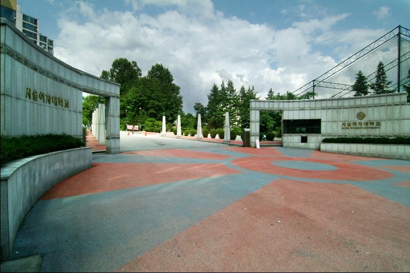서울여자대학교 정문 — 좌우로 나뉜 회색 석재 담장에 한글과 영문 교명이 새겨져 있고, 그 사이 붉은색과 푸른색 무늬가 깔린 넓은 진입 광장 뒤로 나무가 우거져 있다