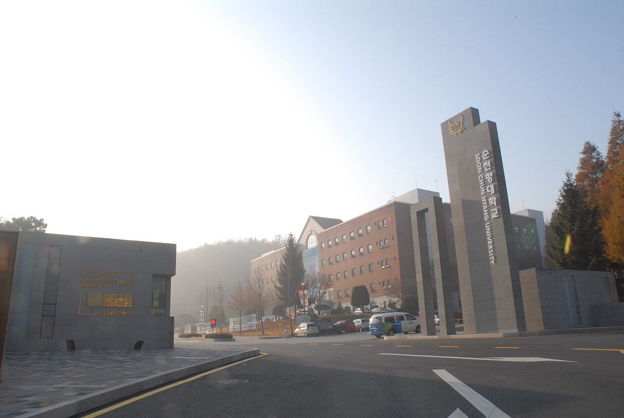 순천향대학교 정문 — 화강암 기둥에 '순천향대학교 SOON CHUN HYANG UNIVERSITY'라고 세로로 새겨져 있고, 왼쪽 낮은 담장에는 'SCH SINCE 1978' 표지가 있다. 뒤로 캠퍼스 건물과 낮은 산이 보인다